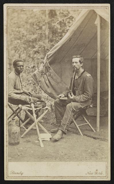 [Unidentified soldier of 23rd New York Infantry Regiment smoking a pipe and unidentified African American man, probably a servant, with sword, sitting in front of tent]