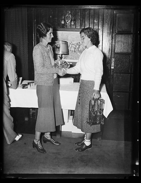 [Two women (Marion McDougall at right?), probably at a sports/golf club (golf balls are on the table), with one woman holding a carrying case with bottles]
