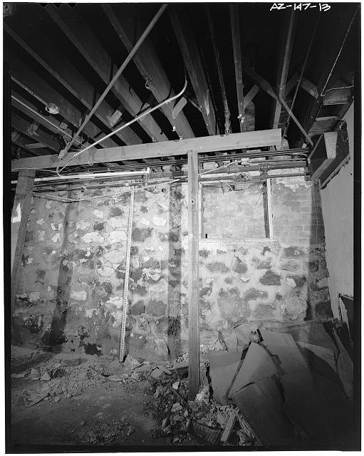 13.  The south segment of the building has a stone basement. The alley wall had a number of areaway windows that are now infilled with bricks. These areaways were subsequently filled with earth, probably when the alley was paved. Here the first-floor joists are seen with a make-shift support beam and column. The basement floor originally was part earth and part wood. Some of the earth floor is now covered with a concrete slab; the wood floor remains. Credit GADA/MRM. - Stroud Building, 31-33 North Central Avenue, Phoenix, Maricopa County, AZ