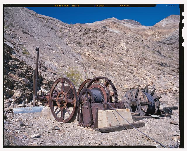 DETAIL VIEW OF WINCHING ENGINE LOCATED AT THE UPPER TRAM TERMINAL, LOOKING NORTHEAST. THE CABLE FROM THIS ENGINE LEADS DOWN INTO THE DEEP RAVINE IN FRONT OF THE UPPER TRAM TERMINAL. IT WAS PROBABLY USED TO DRAG MATERIALS UP TOWARD THE TERMINAL WHEN THE TERMINAL WAS BEING CONSTRUCTED, OR IN TIMES OF TRAMWAY BREAKDOWN. THE DRIVE ENGINE IS IN THE BACKGROUND. TWO LONG OPERATING LEVERS FOR THE ENGINE ARE IN THE CENTER FOREGROUND. AN EXTRA SPOOL OF CABLE IS ON THE GROUND TO THE RIGHT OF THE ENGINE. A WATER PIPELINE STRETCHES ACROSS THE SLOPE IN THE BACKGROUND, CARRYING WATER TO THE UPPER MINES. SEE CA-291-37 FOR IDENTICAL B&W NEGATIVE.  - Keane Wonder Mine, Park Route 4 (Daylight Pass Cutoff), Death Valley Junction, Inyo County, CA
