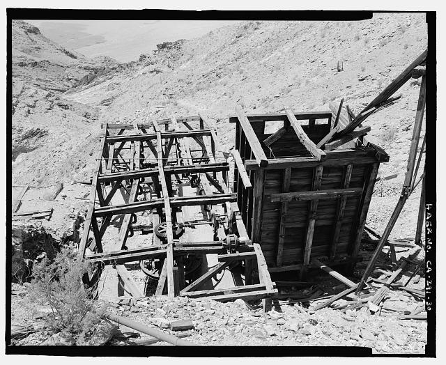 TOP VIEW OF UPPER TRAM TERMINAL, PRIMARY ORE BIN, AND ORE CHUTE,LOOKING SOUTHWEST. TRAM MACHINERY AND GEARS ARE AT LOWER CENTER. A SMALL ELECTRIC MOTOR AT THE REAR LEFT OF THE TERMINAL PROBABLY WAS ADDED AFTER THE ORIGINAL CONSTRUCTION. THE MOVING CABLE OF THE TRAM WAS DRIVEN BY THESE GEARS AND THE LARGE WHEEL UNDERNEATH (SEE CA-291-31 FOR DETAIL). EMPTY TRAM BUCKETS CAME IN FROM THE LEFT, SWINGING AROUND TO THE CHUTES FROM THE ORE BIN TO BE LOADED FOR THE TRIP DOWN TO THE MILL (SEE CA-291-35 FOR DETAIL). THE BREAK OVER TOWER CAN BE SEEN IN THE DISTANCE AT TOP LEFT. THE SUPPORT TOWER BETWEEN THE UPPER TERMINAL AND THE BREAK OVER TOWER IS COLLAPSED.  - Keane Wonder Mine, Park Route 4 (Daylight Pass Cutoff), Death Valley Junction, Inyo County, CA