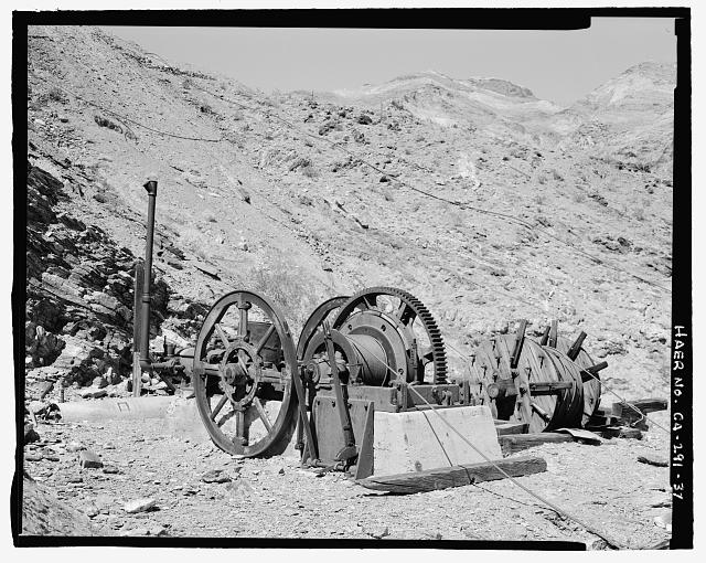 DETAIL VIEW OF WINCHING ENGINE LOCATED AT THE UPPER TRAM TERMINAL LOOKING NORTHEAST. THE CABLE FROM THIS ENGINE LEADS DOWN INTO THE DEEP RAVINE IN FRONT OF THE UPPER TRAM TERMINAL. IT WAS PROBABLY USED TO DRAG MATERIALS UP TOWARD THE TERMINAL WHEN THE TERMINAL WAS BEING CONSTRUCTED, OR IN TIMES OF TRAMWAY BREAK DOWN. THE DRIVE ENGINE IS IN THE BACKGROUND. TWO LONG OPERATING LEVERS FOR THE ENGINE ARE IN THE CENTER FOREGROUND. AN EXTRA SPOOL OF CABLE IS ON THE GROUND TO THE RIGHT OF THE ENGINE. A WATER PIPELINE STRETCHES ACROSS THE SLOPE IN THE BACKGROUND, CARRYING WATER TO THE UPPER MINES. SEE CA-291-52 (CT) FOR IDENTICAL COLOR TRANSPARENCY.  - Keane Wonder Mine, Park Route 4 (Daylight Pass Cutoff), Death Valley Junction, Inyo County, CA