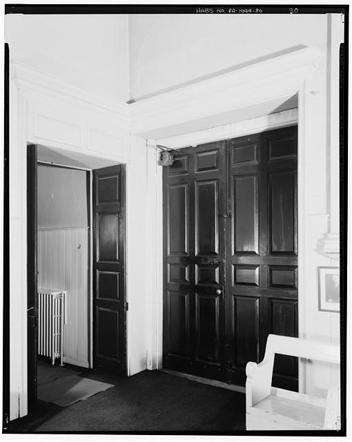 30.  ROOM A (WEST ROOM), SOUTHEAST CORNER, LOOKING SOUTHEAST. The south exterior doors are at right. Note the door closer pulley and weight box still in place. The doorway at left leads to the Main Meeting Room. The overdoor paneling probably was added c.1892. Note the capped gas pipe to the right of the exterior doors. - Twelfth Street Meeting House, 20 South Twelfth Street, Philadelphia, Philadelphia County, PA